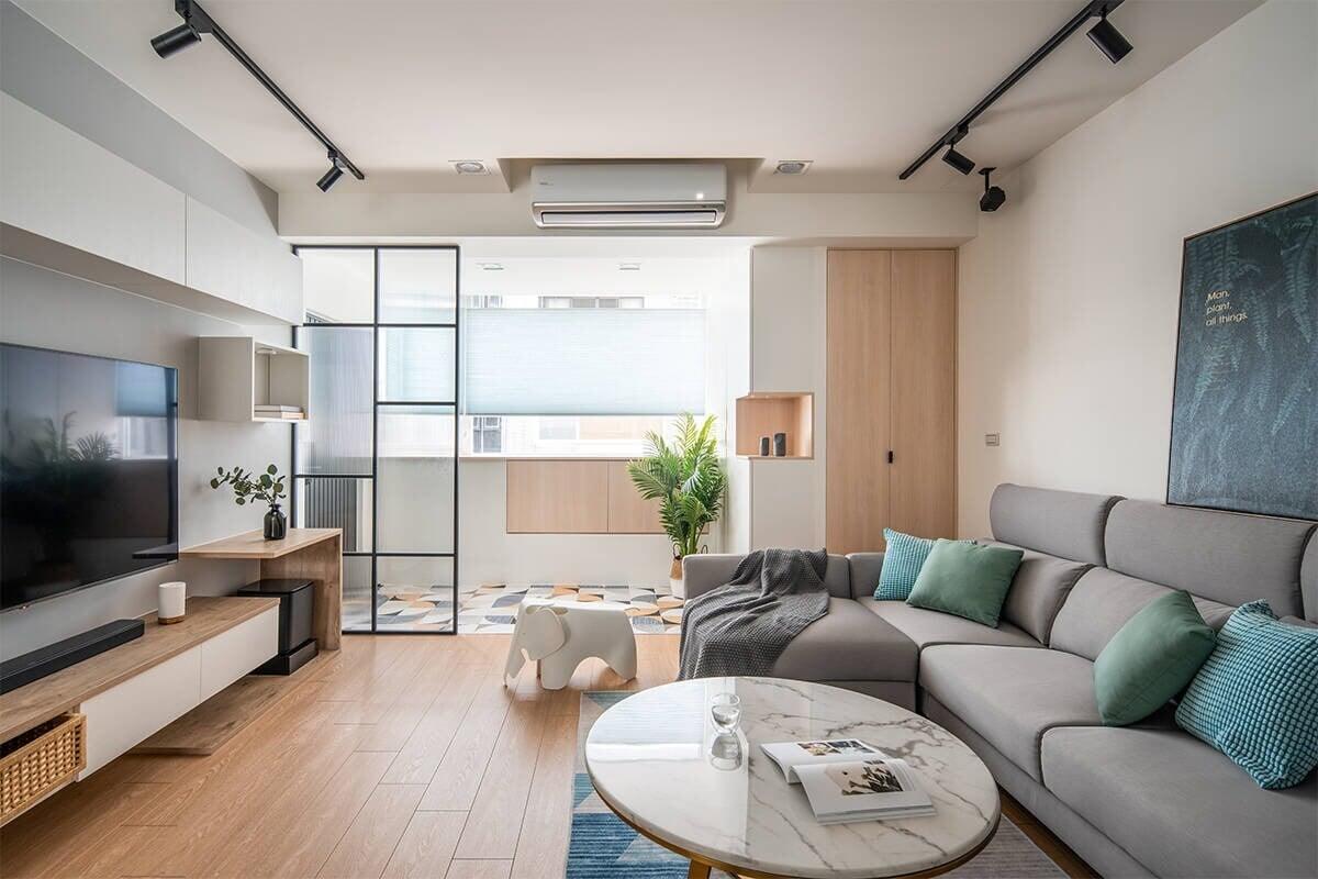 Phòng khách hiện đại với sofa xám, bàn tròn, và cửa kính, trang trí cây xanh và đèn chiếu ánh sáng.