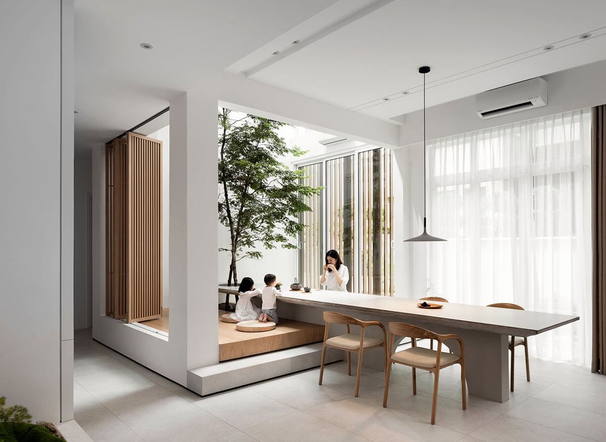 Phòng ăn hiện đại với nội thất gỗ, có cây xanh và mọi người đang ngồi thư giãn uống trà gần cửa sổ lớn.