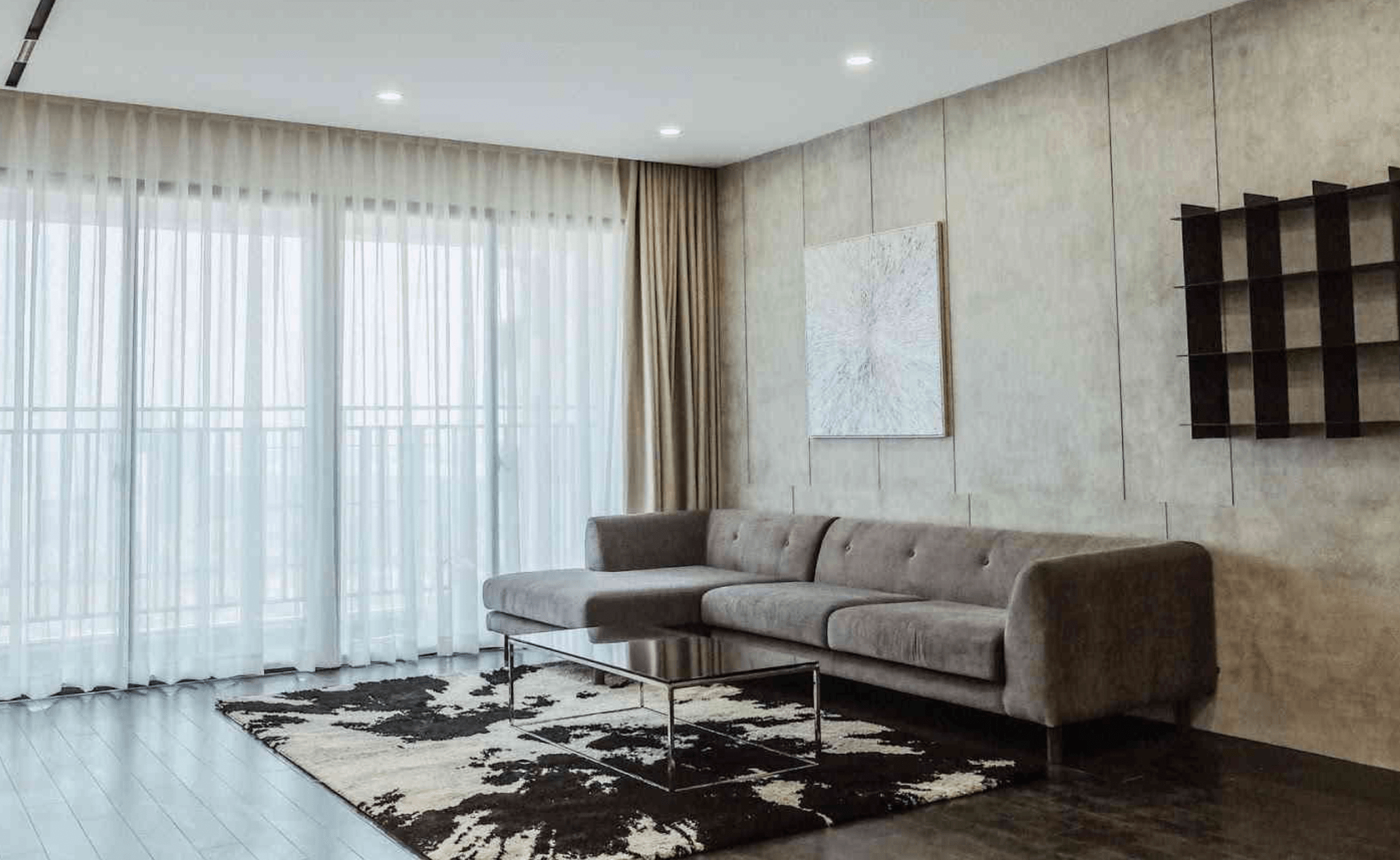 Phòng khách hiện đại với ghế sofa xám, thảm họa tiết, bàn kính và rèm cửa lớn, ánh sáng thiên nhiên tràn ngập.