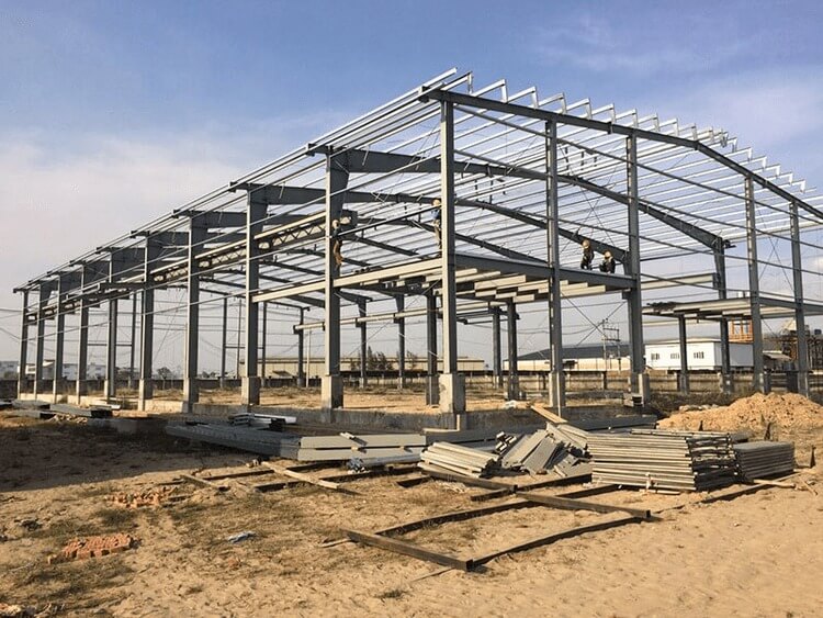 Công trường xây dựng nhà xưởng với khung thép lớn, vật liệu nằm rải rác trên mặt đất.