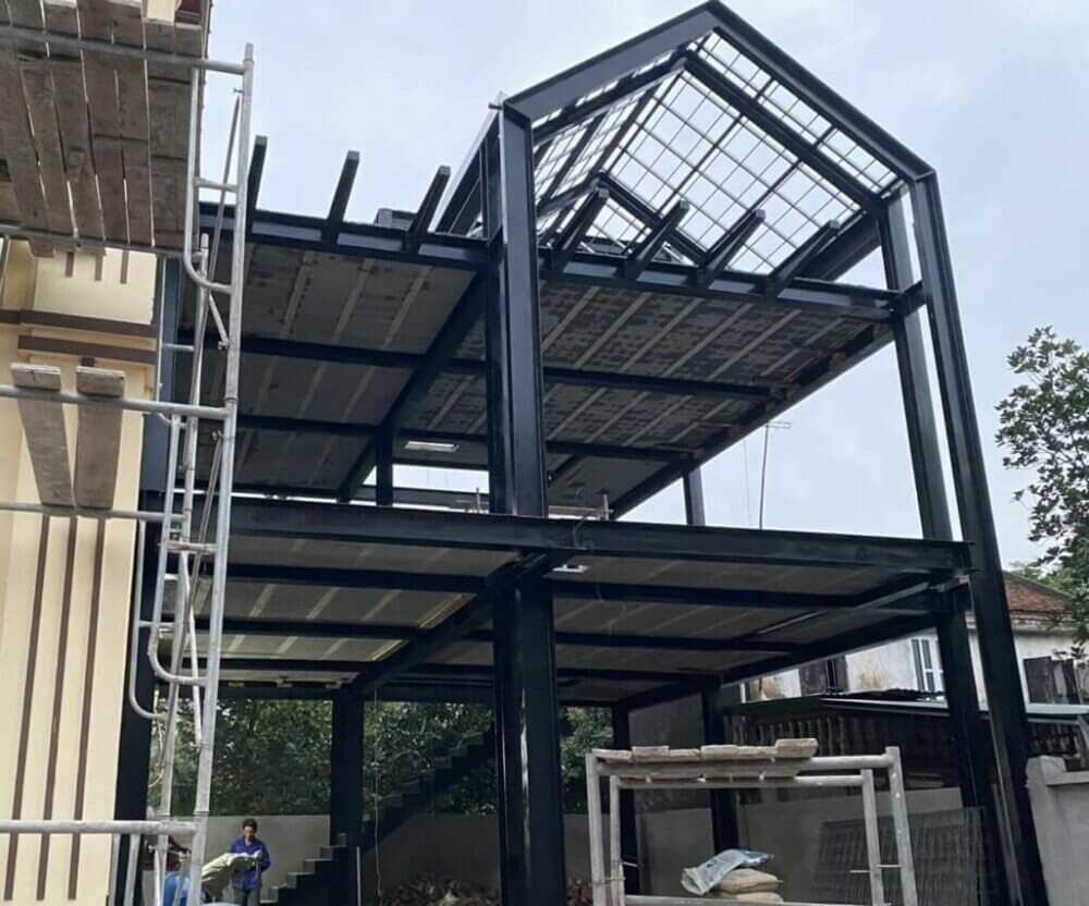 Khung nhà thép đang xây dựng, giàn giáo dựng phía bên trái, bầu trời trong xanh.