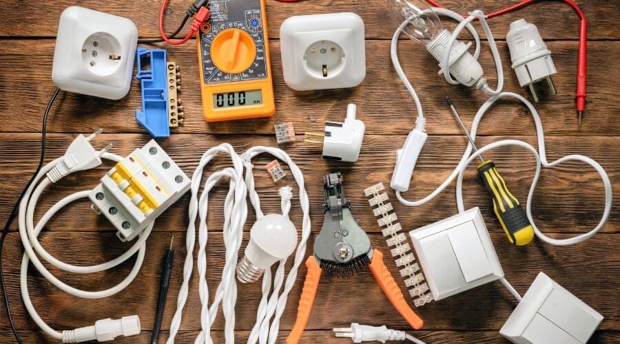 Dụng cụ và linh kiện điện trên bàn gỗ bao gồm ổ cắm, công tắc, đèn, vít, kềm và đồng hồ đo điện.