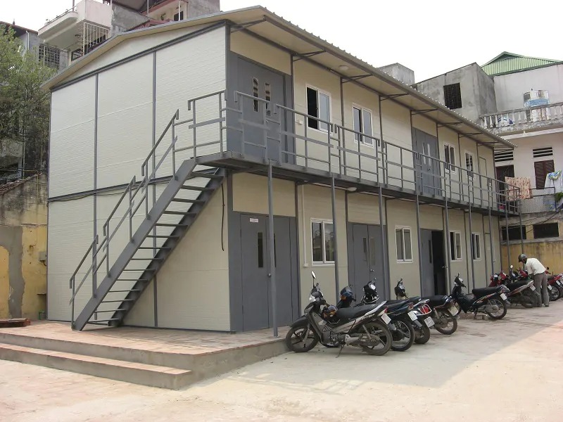 Nhà tiền chế hai tầng với cầu thang bên ngoài. Xe máy đậu trước tòa nhà trong một khu dân cư.