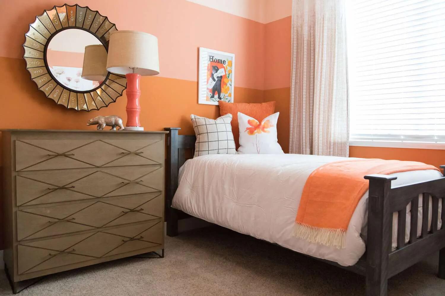 Phòng ngủ trẻ em với giường trắng và chăn cam, gương tròn và đèn bàn trang trí.