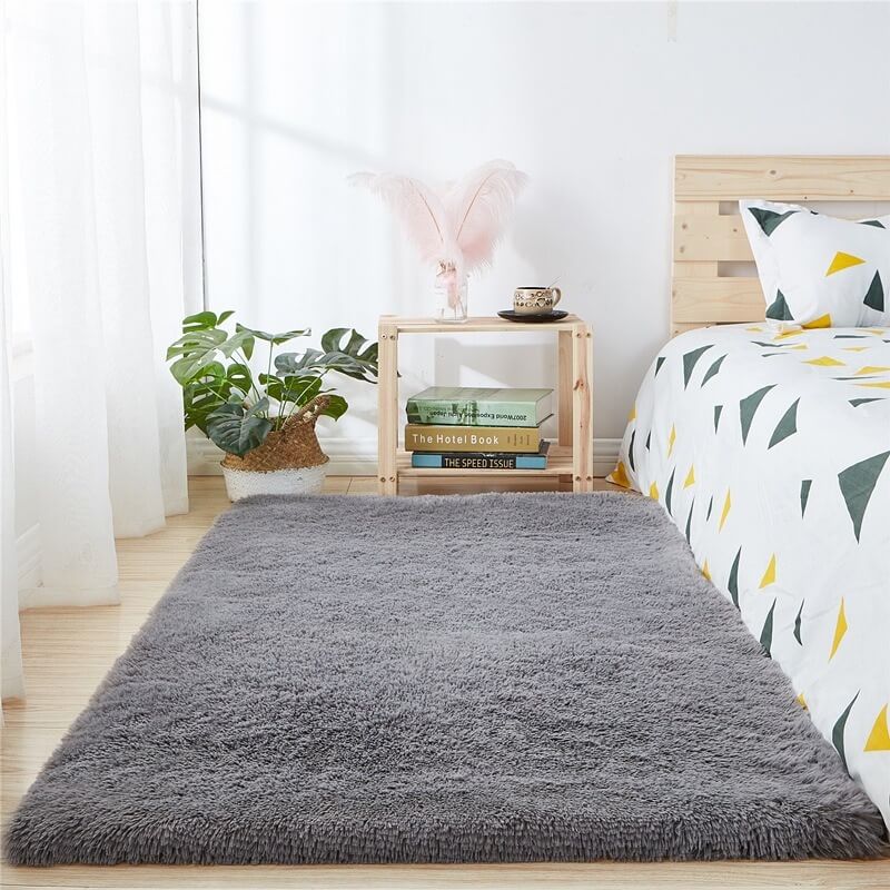 Phòng ngủ sáng sủa với thảm lông màu xám, giường gỗ, gối tam giác, và cây xanh trang trí trên kệ.