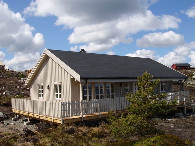 Ngôi nhà gỗ với hàng rào trắng, nằm trên cánh đồng vắng với bầu trời xanh và mây trắng.