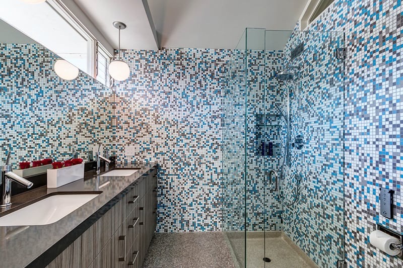 Phòng tắm hiện đại với gạch mosaic xanh, vách kính, bồn rửa đôi, và đèn chiếu sáng.