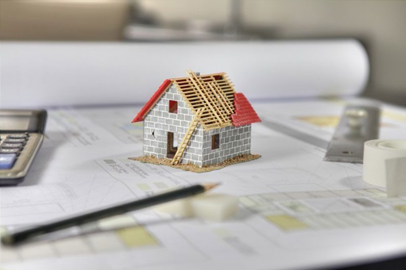 Mô hình nhà nhỏ trên bản vẽ kiến trúc, cùng bút chì và máy tính, biểu thị kế hoạch xây dựng.