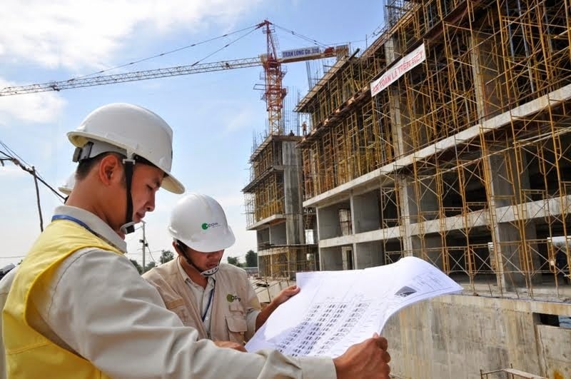 Hai kỹ sư xây dựng đang xem bản vẽ trước công trình dang dở, có cần cẩu và giàn giáo phía nền trời xanh.