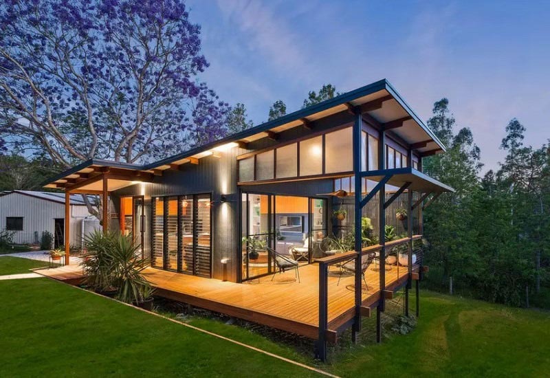 Nhà gỗ hiện đại với cửa kính lớn, được bao quanh bởi cây cối xanh mát.