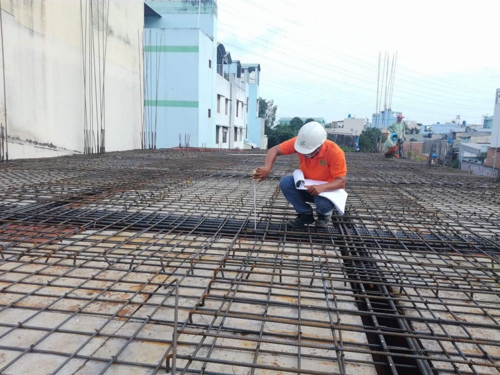 Kỹ sư giám sát công trường xây dựng, kiểm tra lưới thép trên mái, đội mũ bảo hộ, nền trời trong xanh.