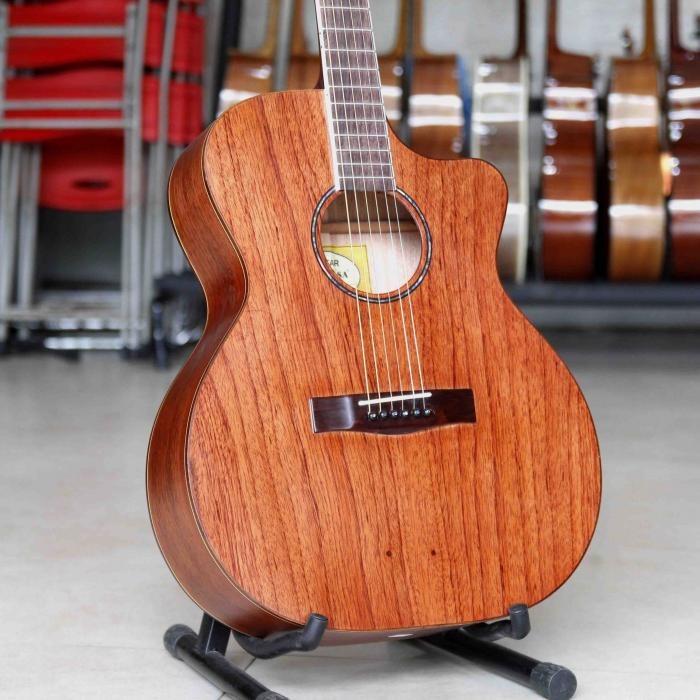 Guitar acoustic gỗ tự nhiên trên giá đỡ, nền có nhiều guitar khác trong cửa hàng nhạc cụ.