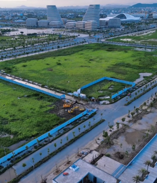 Khu đất đang xây dựng với máy xúc, xung quanh là hàng rào xanh và tòa nhà hiện đại ở phía xa.