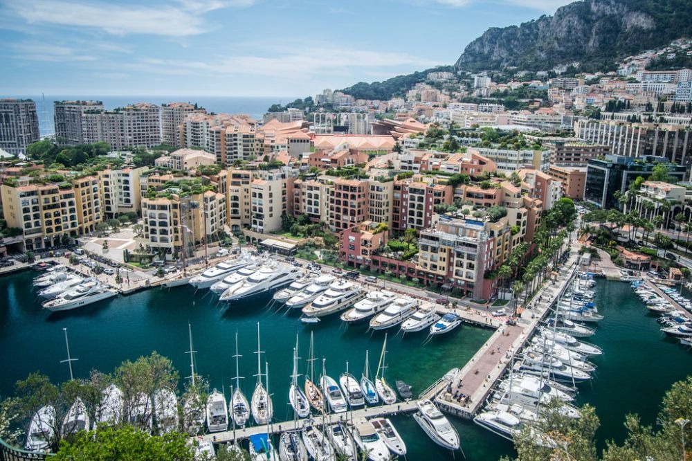 Bến cảng Monaco với du thuyền sang trọng, tòa nhà đẹp và núi non phía sau. Trời xanh tạo không khí thư giãn.