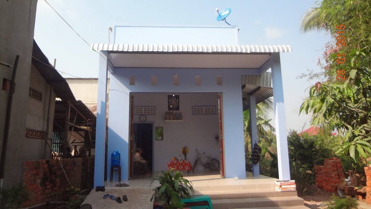 Nhà cấp 4 sơn màu xanh dương nhạt có mái tôn, cây xanh xung quanh, một người ngồi bên trong cửa.