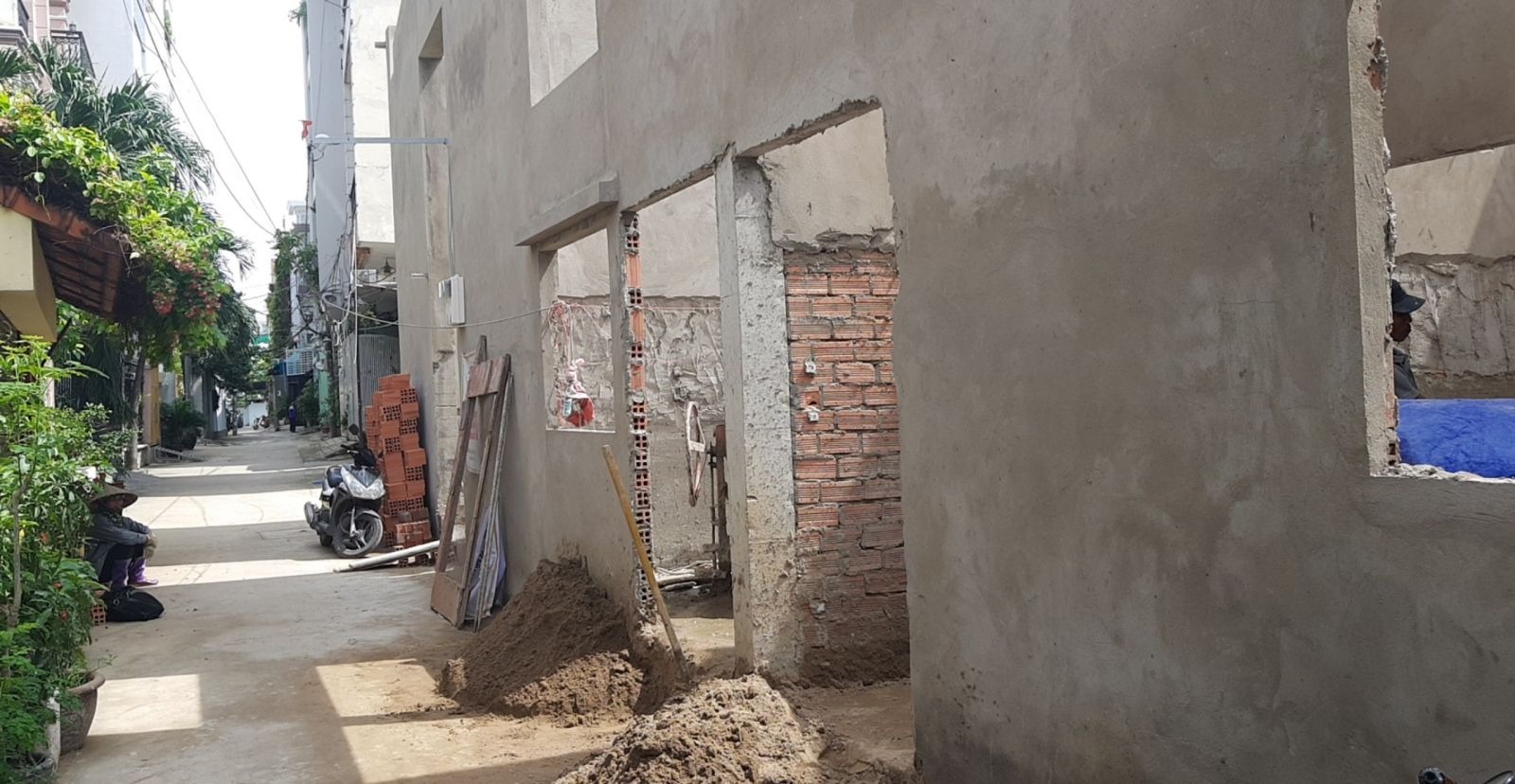 Nhà đang xây dựng ở khu phố nhỏ với xe máy và cây xanh xung quanh. Công trường có gạch và cát.