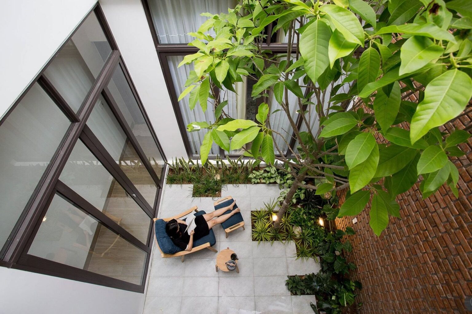 Người phụ nữ thư giãn trên ghế ngoài trời giữa khu vườn xanh, trong một không gian hiện đại với cửa kính lớn.