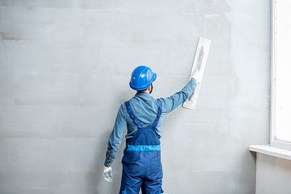 Người thợ xây đang trát tường với mũ bảo hộ xanh, tạo bề mặt nhẵn cho bức tường trong nhà.