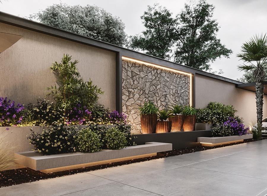 Tường đá và khu vườn cảnh quan với cây xanh và hoa tím, ánh sáng trang trí ban đêm.