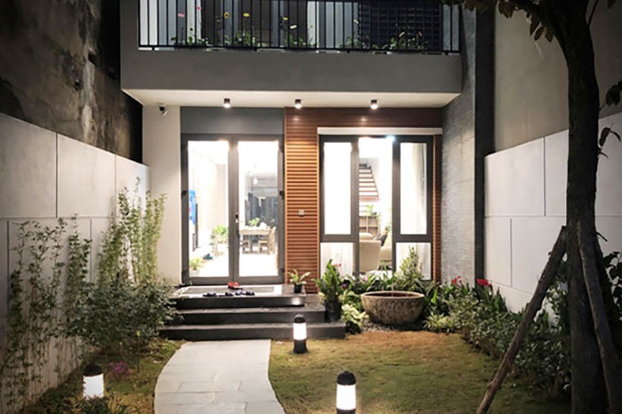 Mặt tiền nhà hiện đại ban đêm với ánh sáng ấm cúng, cây xanh trang trí và lối đi lát đá dẫn vào cửa chính.