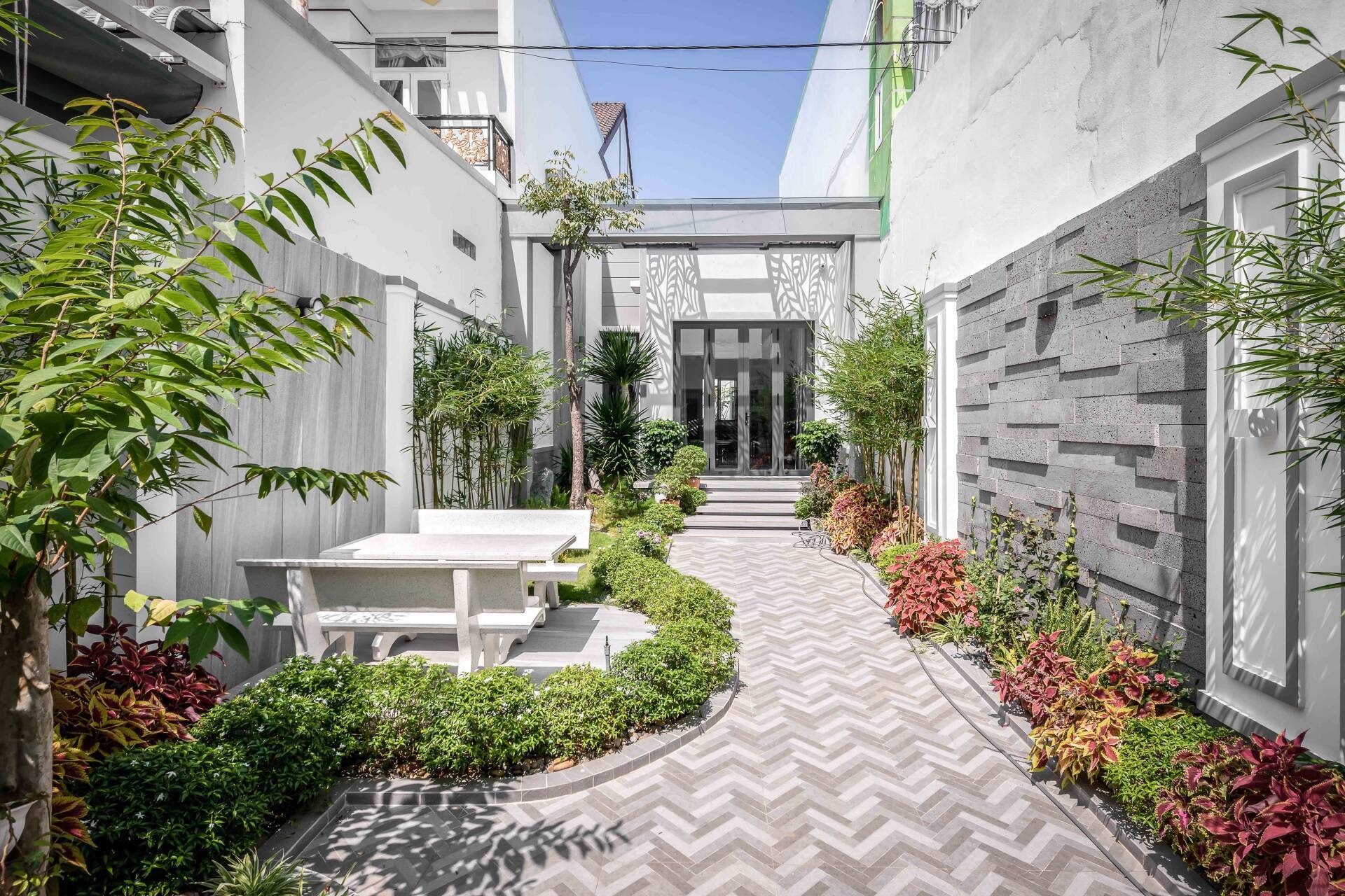 Lối đi sân vườn rộng lát gạch, hai bên trồng cây xanh và bàn ghế đá, tạo không gian thư giãn ngoài trời.