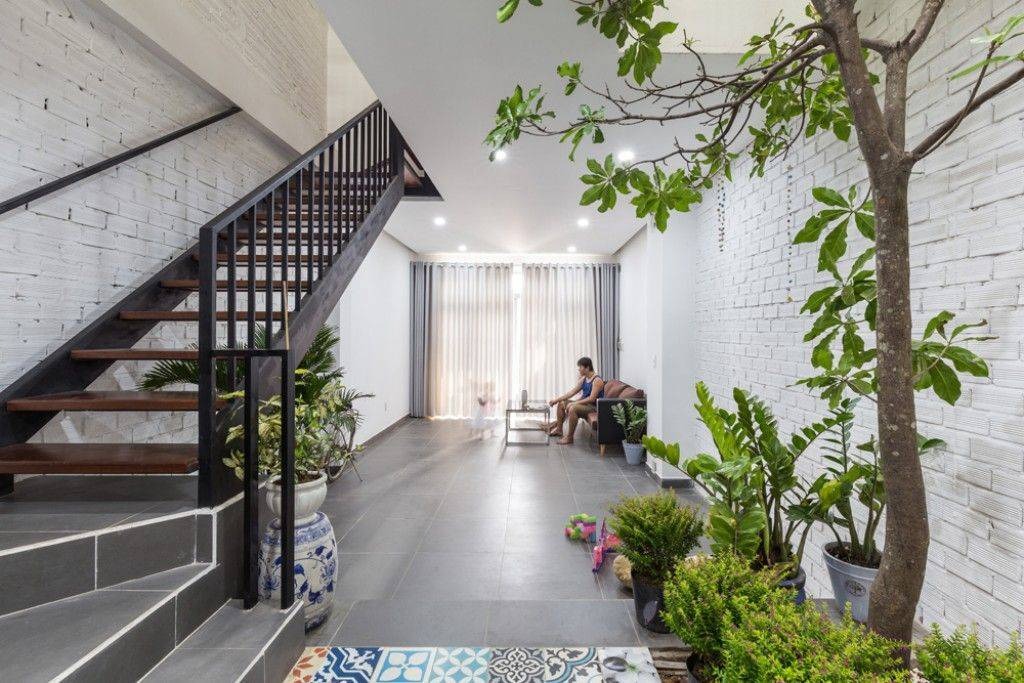 Cầu thang gỗ, cây cảnh, và người ngồi trong phòng khách hiện đại, sáng sủa với tường gạch trắng.