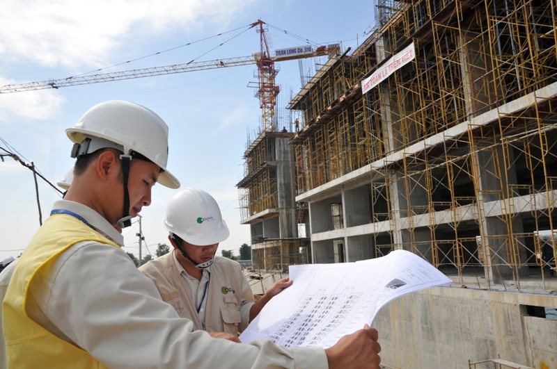 Kỹ sư kiểm tra bản vẽ tại công trường xây dựng với dàn giáo, cần cẩu và tòa nhà đang thi công.