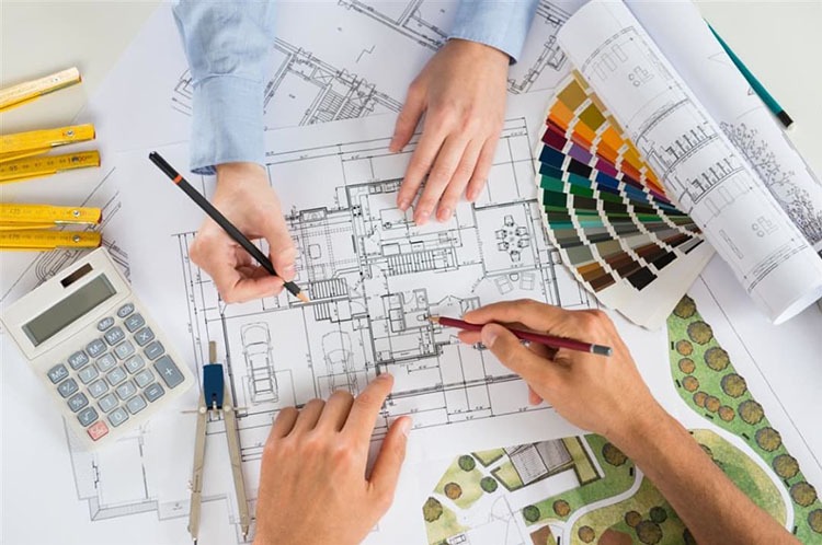 Bản vẽ kiến trúc với tay người, thước kẻ, bảng màu và máy tính, thể hiện quá trình thiết kế nội thất.
