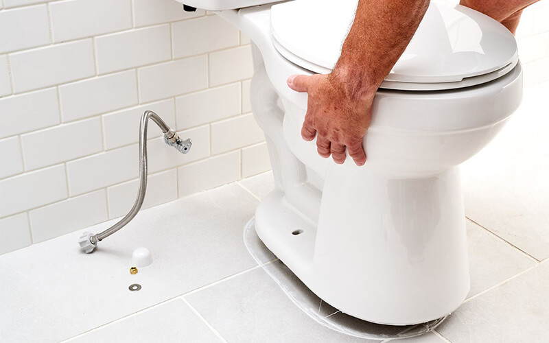 Người đàn ông lắp đặt bồn cầu trong phòng tắm lát gạch trắng, dây dẫn nước và vít lắp đặt bên cạnh.