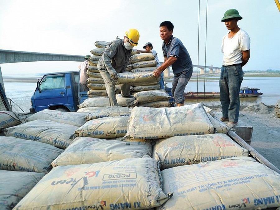 Công nhân đang vận chuyển bao xi măng trên công trường xây dựng gần sông, với xe tải ở phía sau.