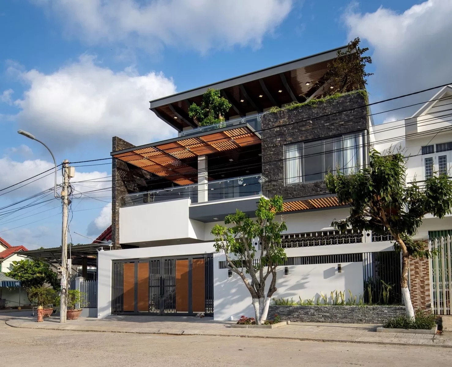 Nhà phố hiện đại, hai tầng với sân thượng, thiết kế mái che gỗ và cây xanh, dưới bầu trời xanh mây trắng.