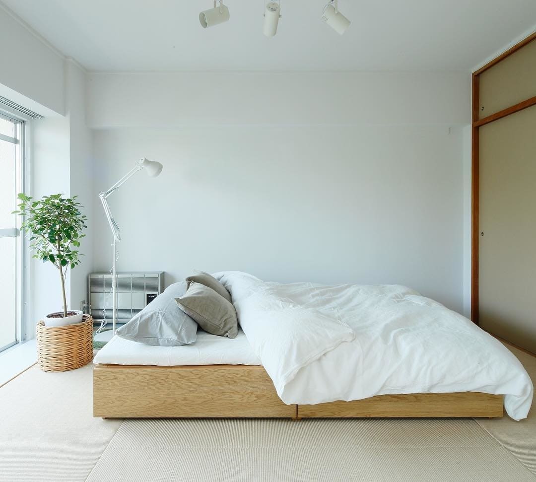 Phòng ngủ tối giản với giường gỗ, chăn trắng, đèn đọc sách và cây xanh gần cửa sổ lớn.