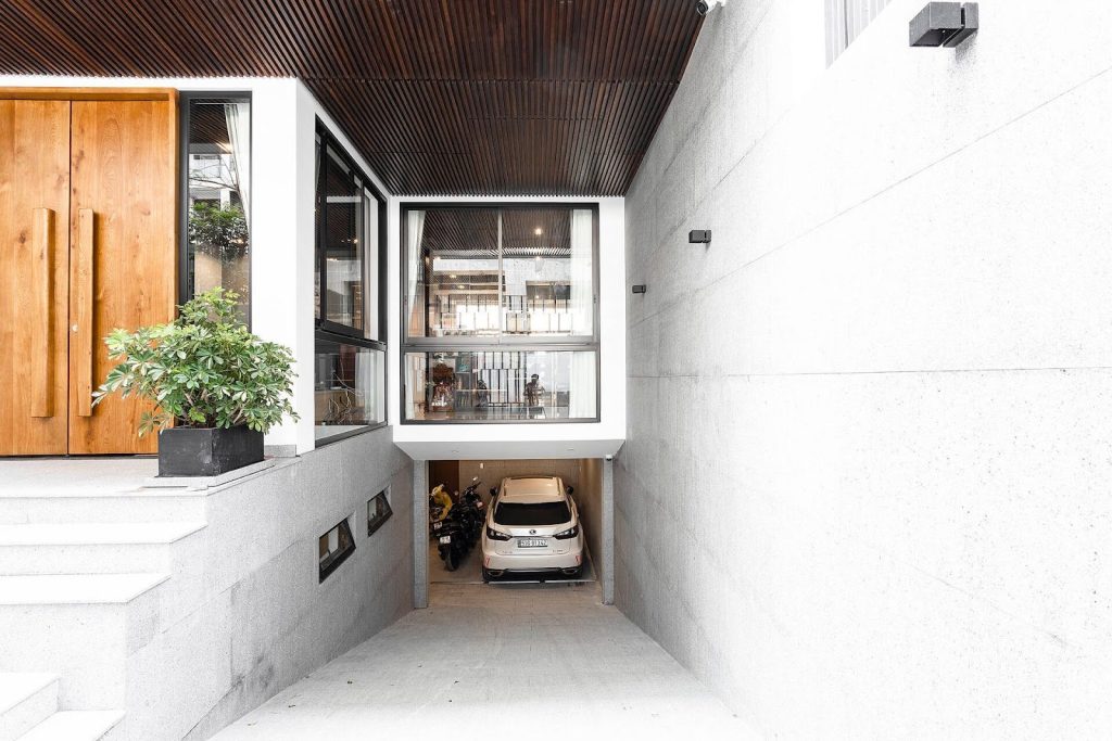 Lối vào gara ngôi nhà hiện đại với cửa gỗ, cây xanh, xe hơi và thiết kế tối giản.