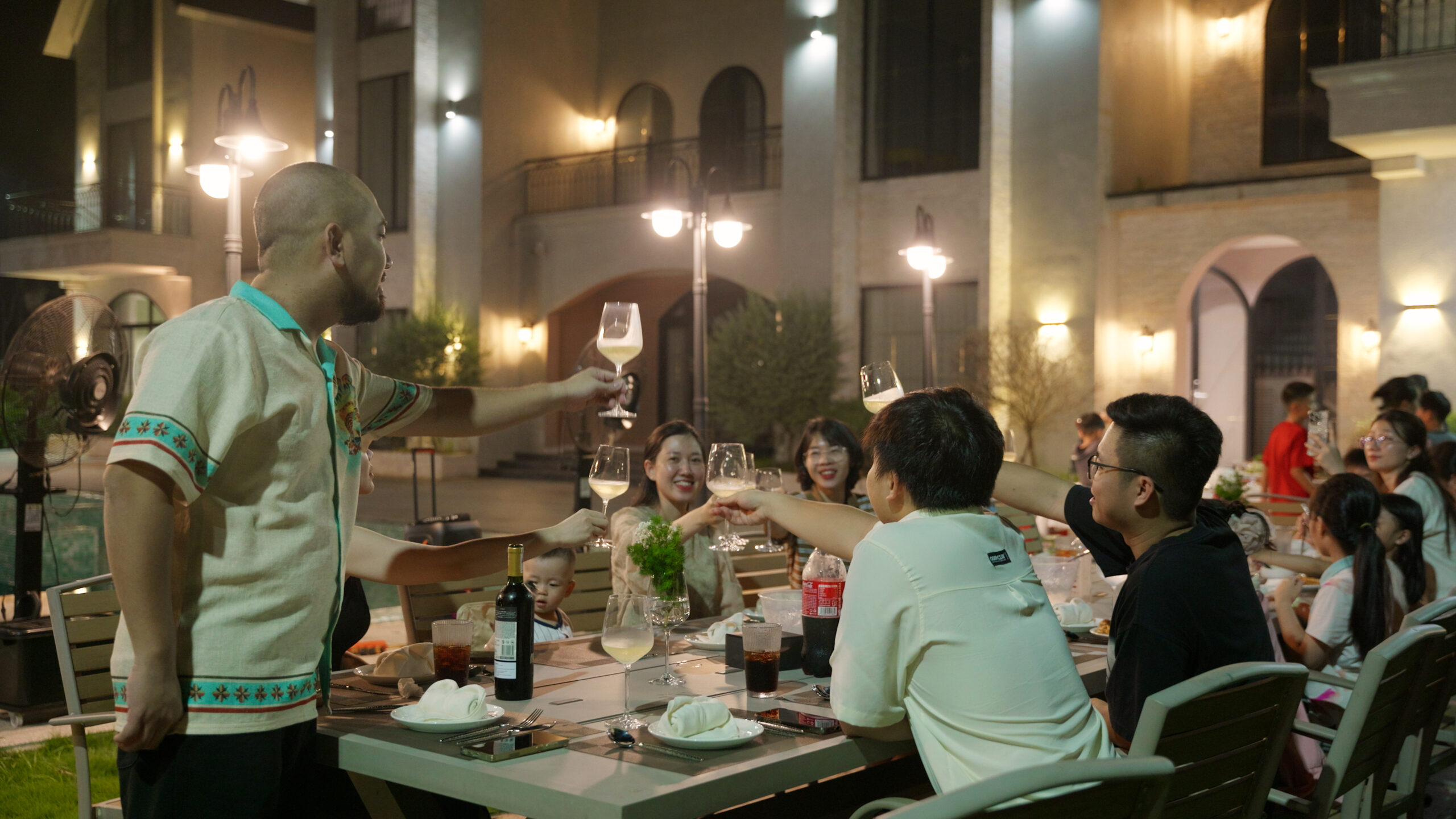 11-scaled-1 Người lớn và trẻ em nâng ly ăn mừng ngoài trời vào buổi tối. Không khí vui vẻ tại một bữa tiệc gia đình.