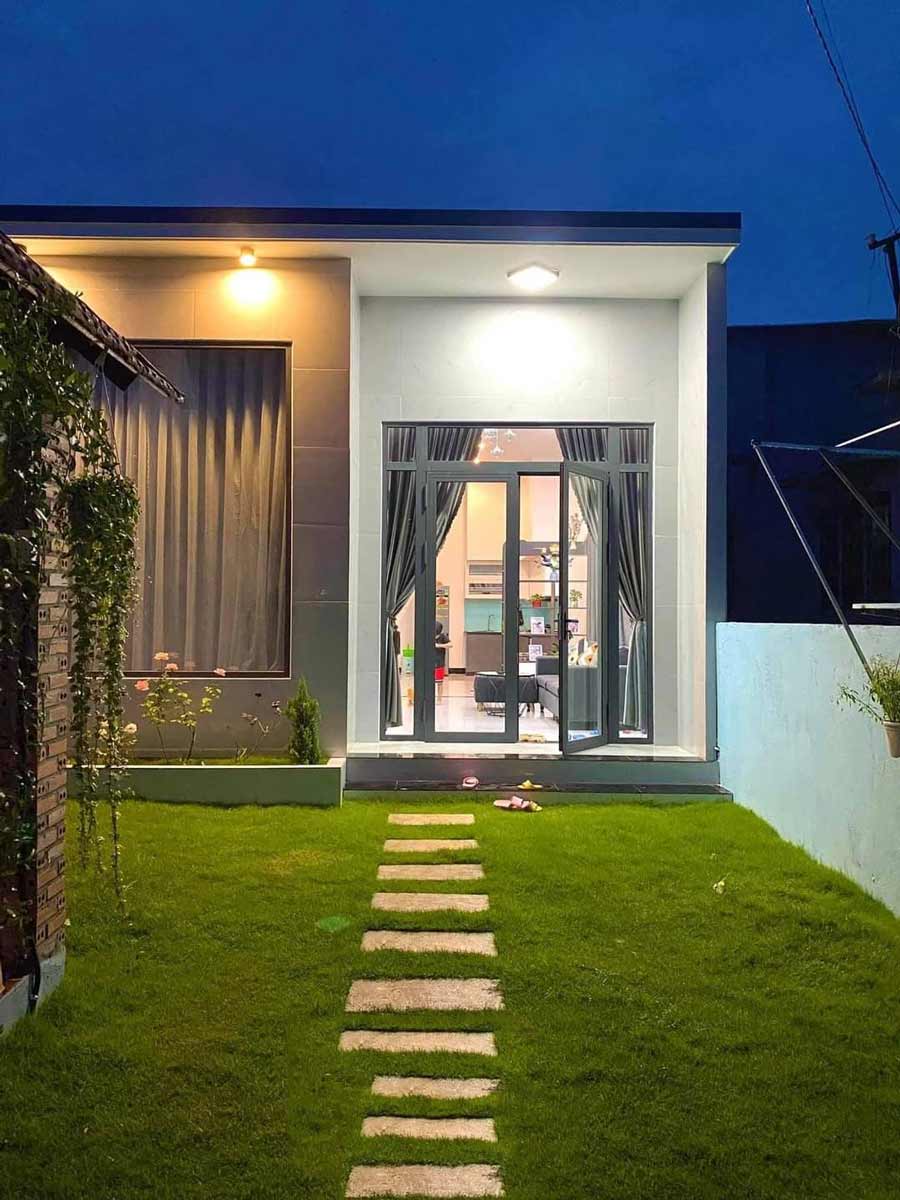 Ngôi nhà một tầng rực sáng ban đêm, lối đi lát đá len qua vườn cỏ xanh, cửa kính lớn dẫn vào phòng khách.