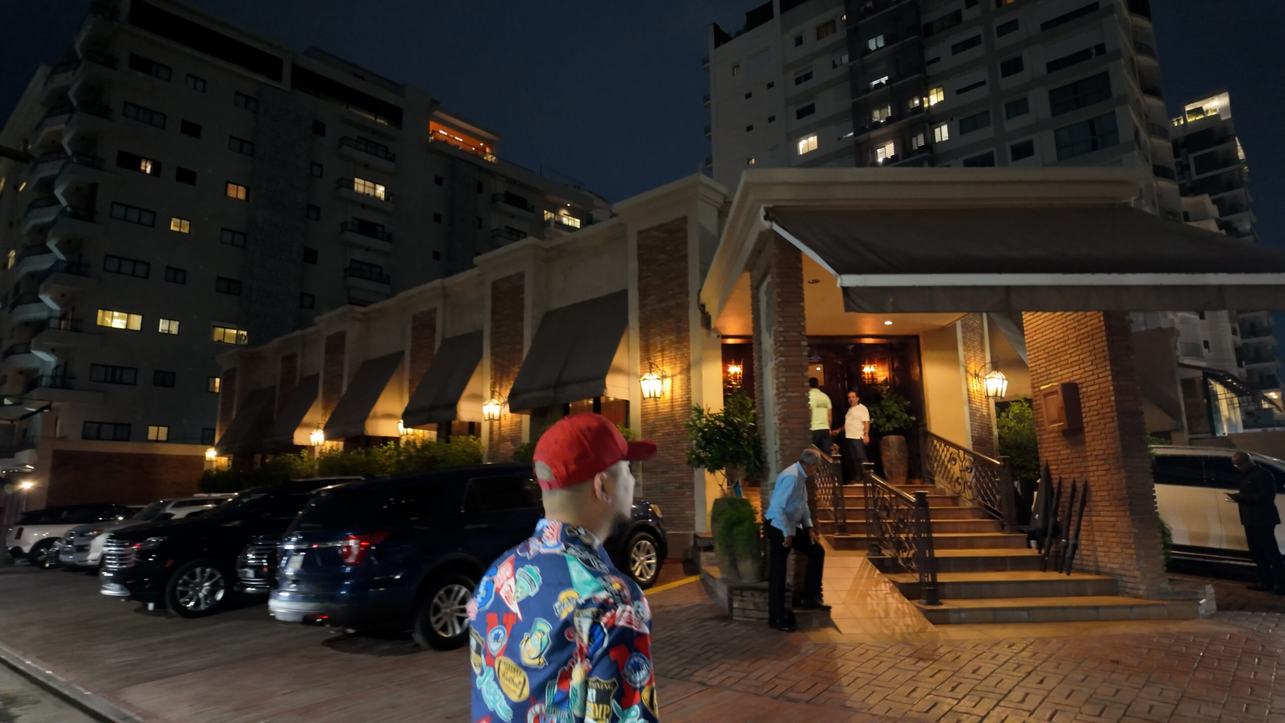 13-1-1-scaled-1 Người đàn ông đội mũ đỏ đang bước vào một tòa nhà lớn vào buổi tối, bên cạnh là hàng loạt xe ô tô đỗ.