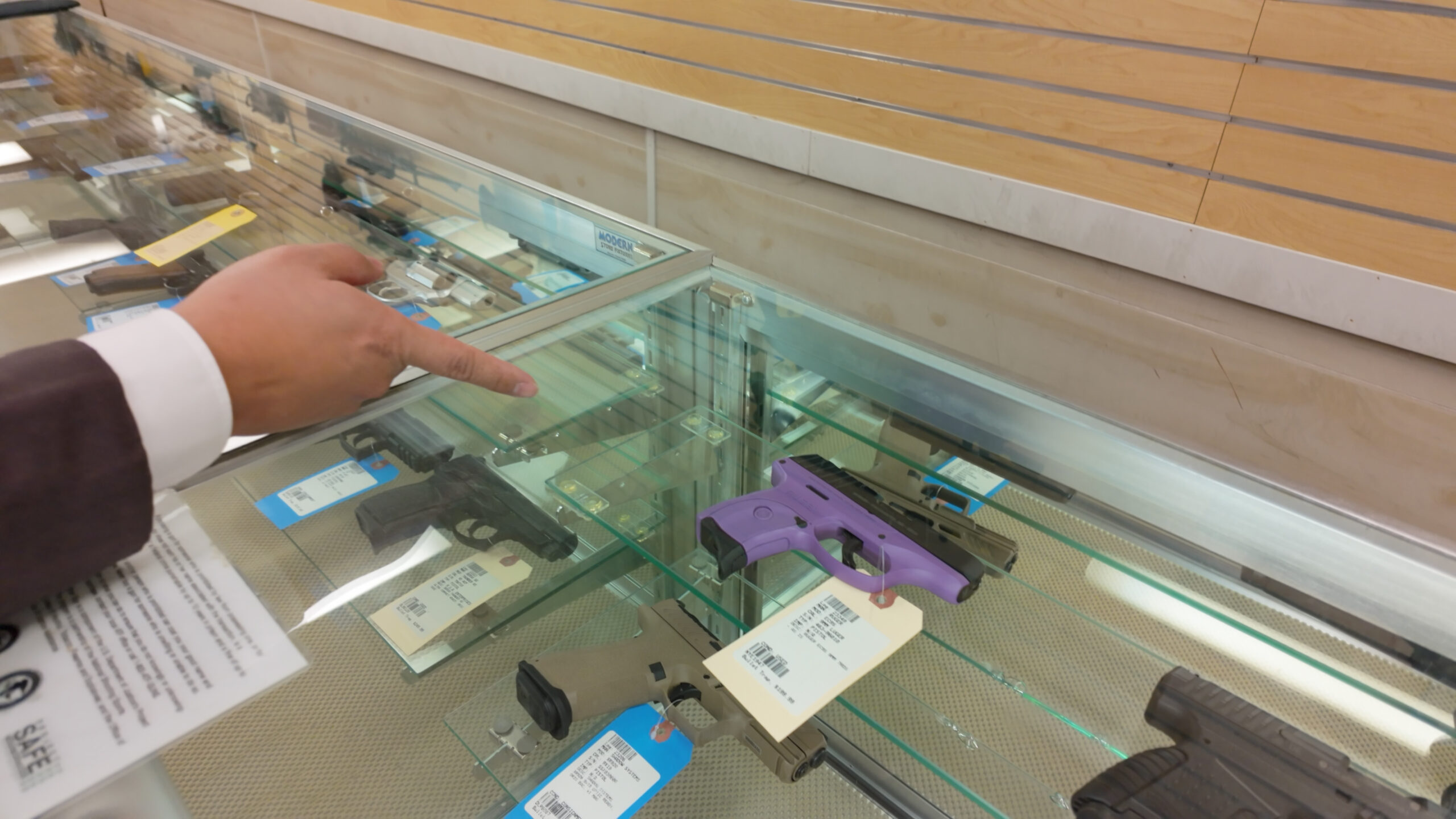 Người chỉ tay vào súng trưng bày trong tủ kính, có súng màu tím và nâu, tại cửa hàng vũ khí.