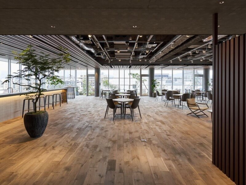 Không gian văn phòng mở với sàn gỗ và cây xanh, bàn ghế hiện đại gần cửa sổ lớn.