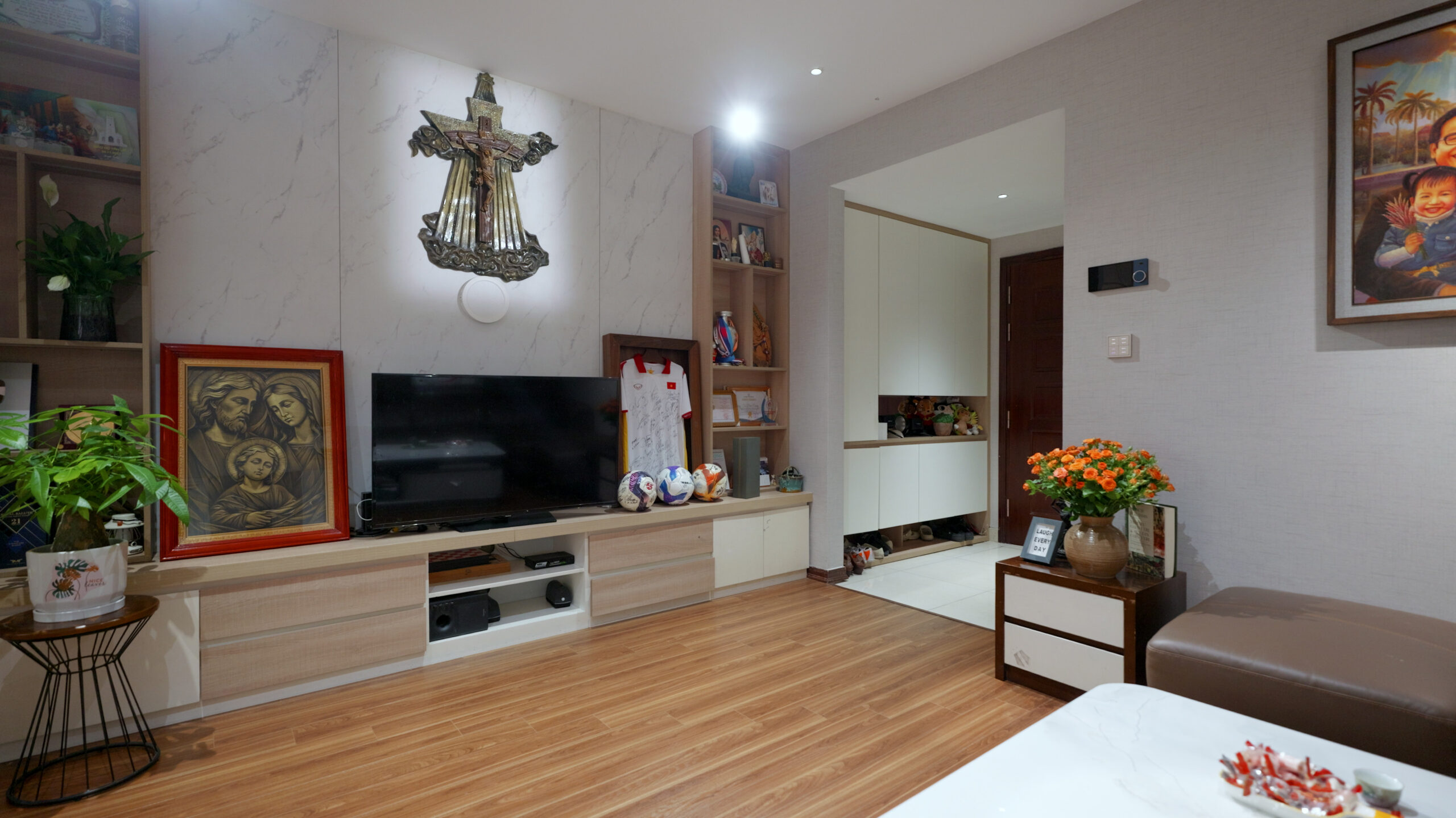 Phòng khách ấm cúng với TV, tranh, bóng đá và cây xanh, kết hợp nội thất gỗ sang trọng.