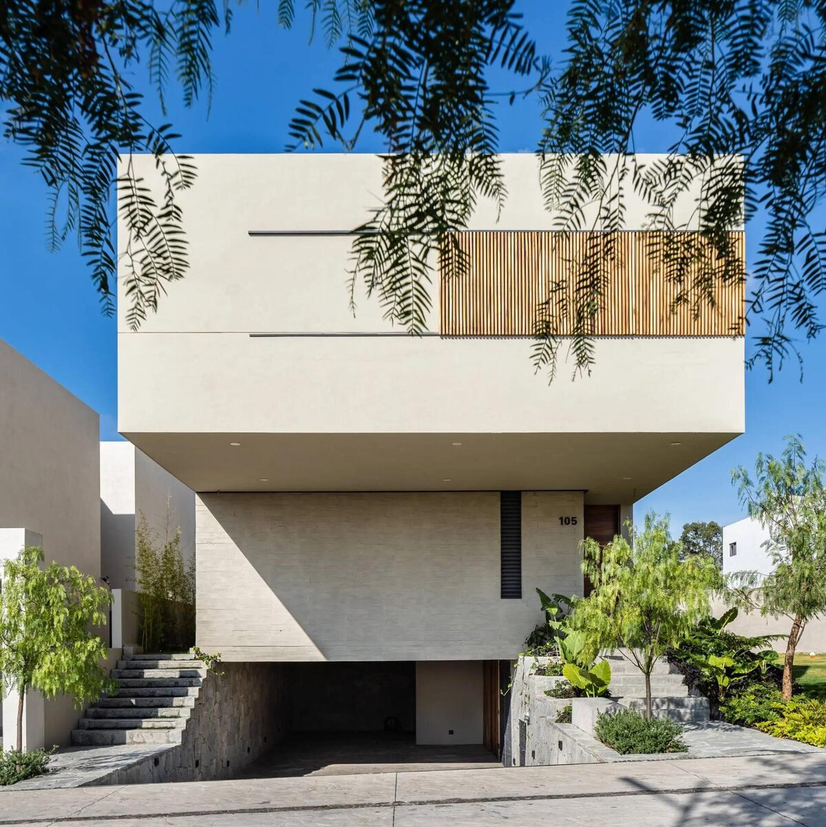 Ngôi nhà hiện đại với thiết kế tối giản, tông màu be, cây xanh bao quanh, nổi bật dưới bầu trời xanh.