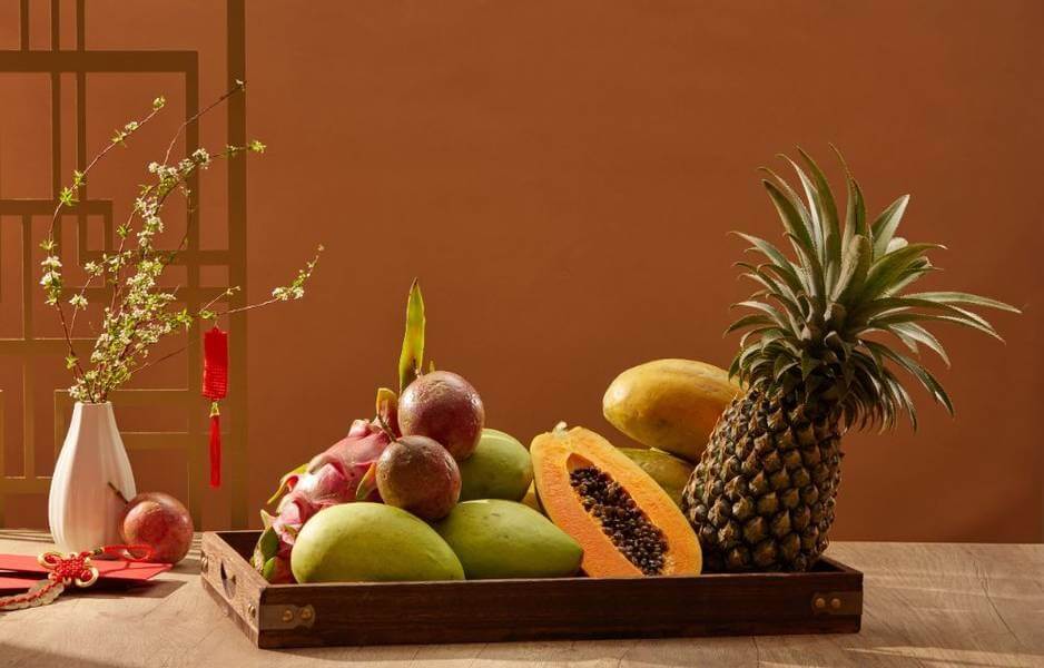 Trái cây nhiệt đới gồm dứa, đu đủ, xoài và thanh long trong khay gỗ, cạnh bình hoa mai trang trí Tết.