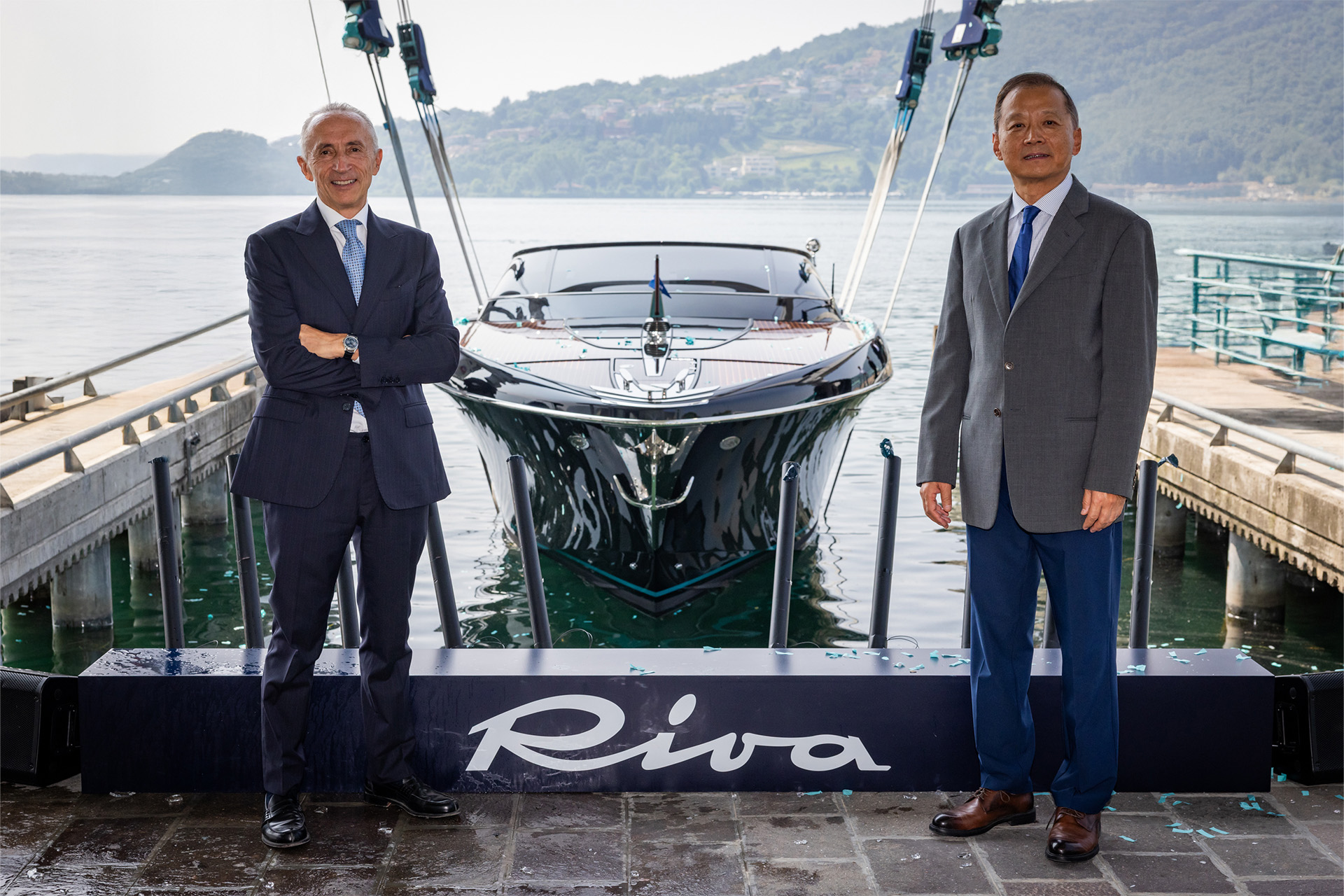 Hai doanh nhân đứng trước du thuyền Riva bên hồ nước, mặc vest lịch sự, nền là núi non.
