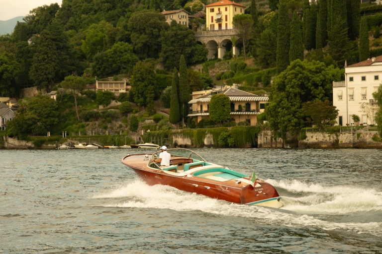 Thuyền máy sang trọng chạy trên hồ cạnh cảnh quan nhà cửa và cây xanh tươi tốt.