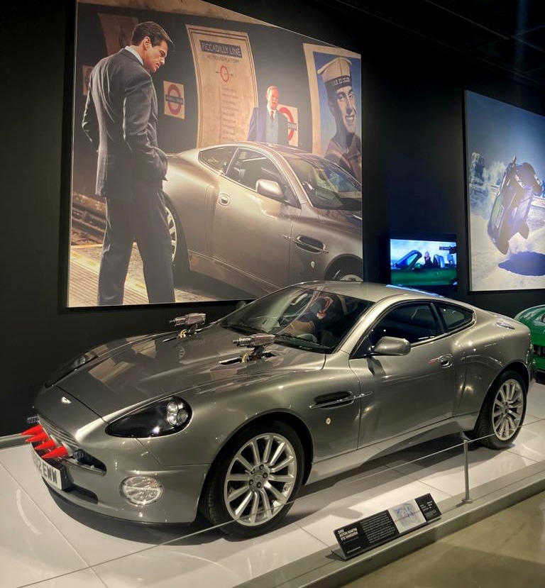 210925-bond-in-motion-los-angele Xe Aston Martin màu bạc trưng bày, nền là áp phích phim có người đàn ông đứng cạnh xe tương tự.