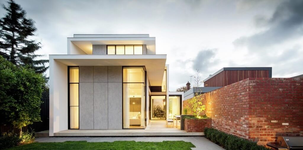 Nhà hiện đại tối giản với cửa kính lớn và gạch đỏ nổi bật, xung quanh là cây cối xanh tươi.