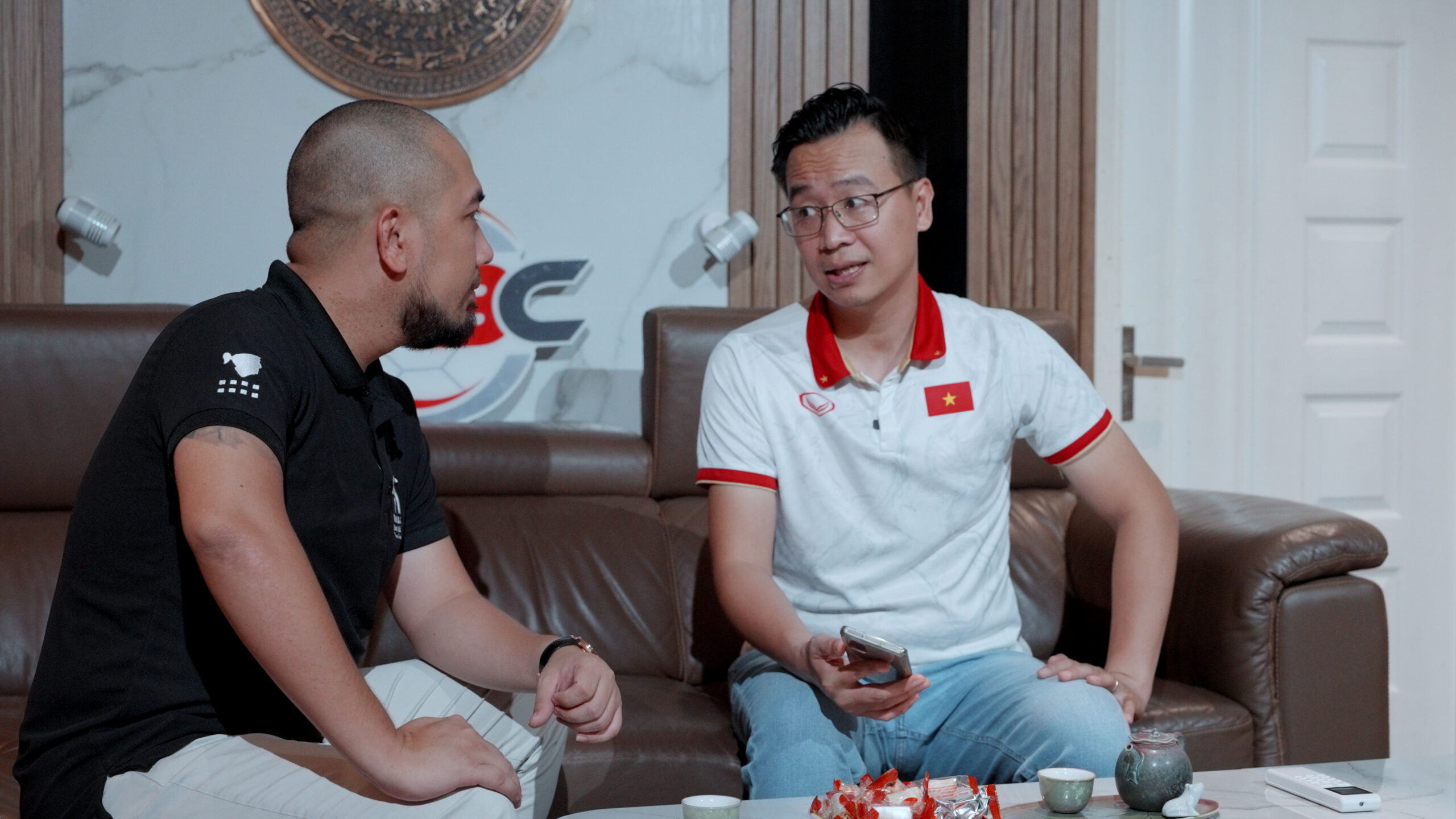 Hai người đàn ông ngồi trên ghế sofa, thảo luận, một người cầm điện thoại.