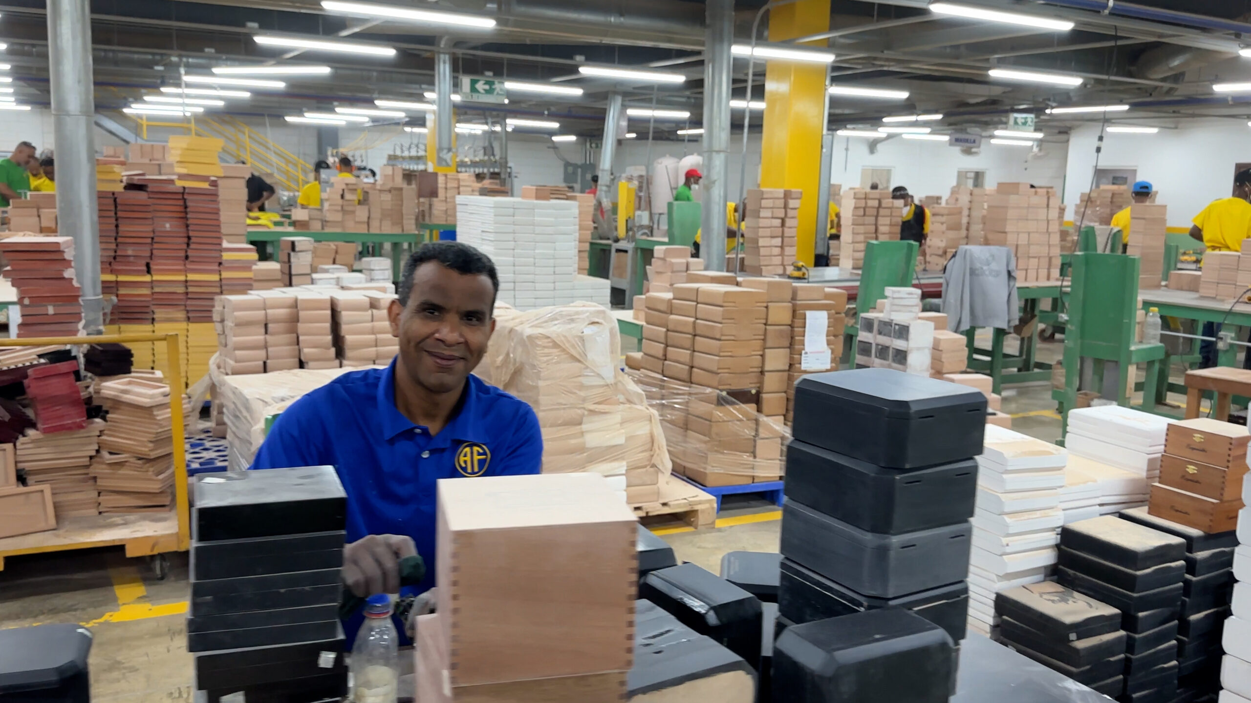 Người đàn ông trong xưởng làm việc với nhiều thùng gỗ xung quanh, ánh sáng đèn trần sáng.