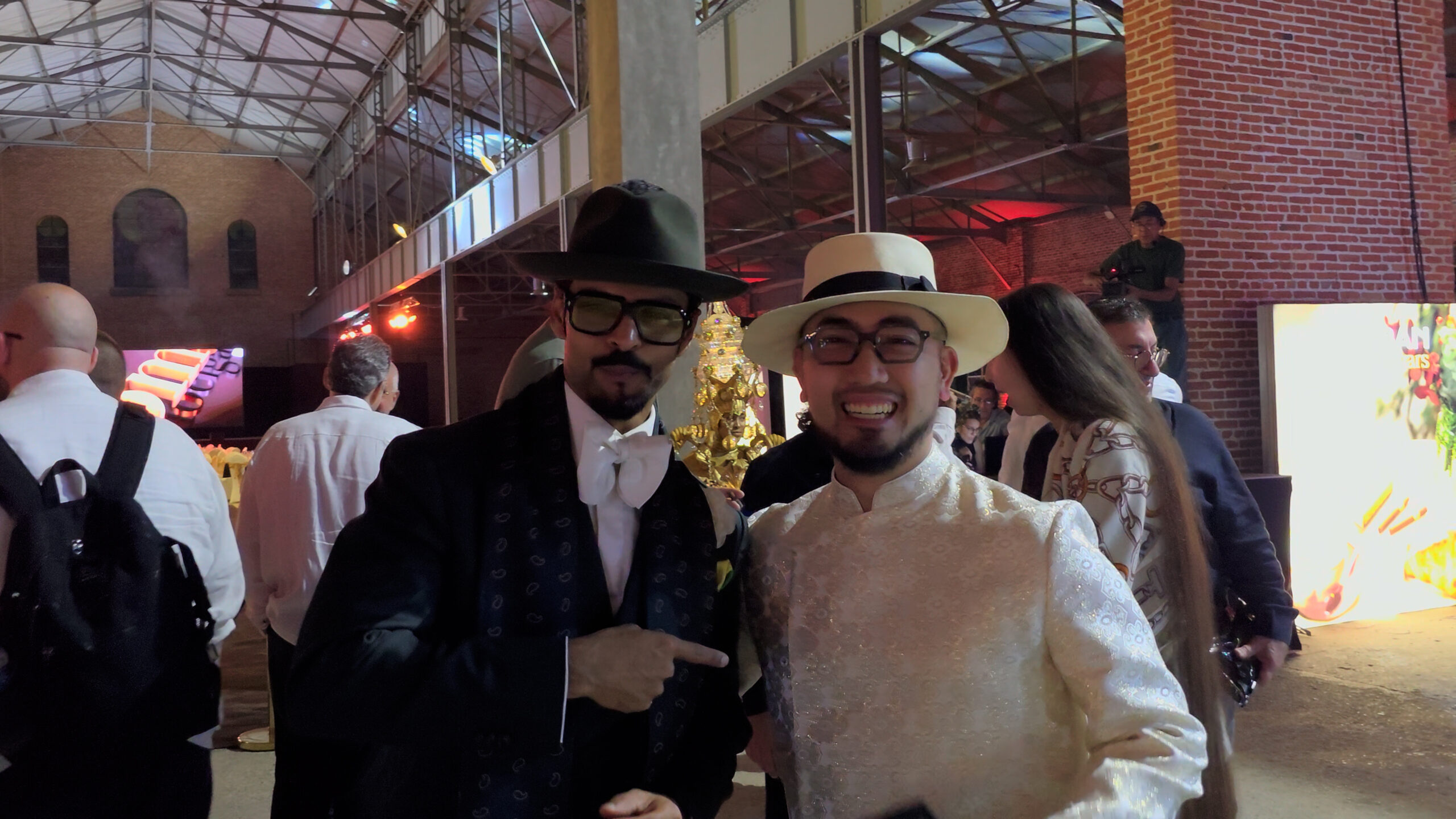 26-scaled-1 Hai người đàn ông đội mũ phong cách đứng trong sự kiện trong nhà với đám đông phía sau.