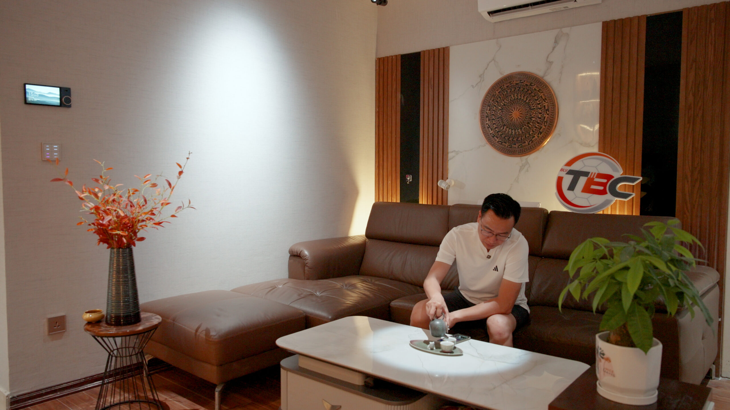 28-scaled-1 Người đàn ông ngồi trên sofa trong phòng khách hiện đại, bên cạnh là bình hoa và cây xanh.