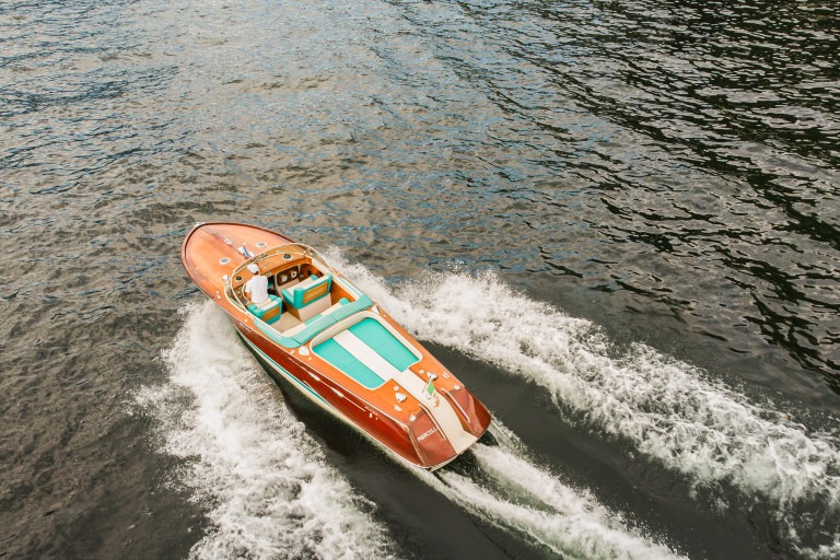 Thuyền máy màu nâu cam chạy trên mặt nước tạo sóng trắng, khung cảnh từ trên cao.